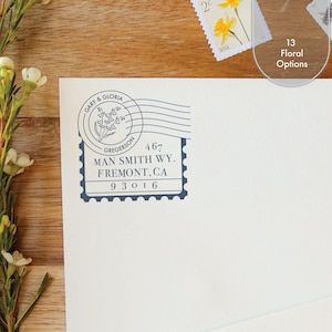 Peut inclure: Une enveloppe blanche avec un tampon à l'encre bleu foncé portant le texte GARY & GLORIA GREGERSON, 467 MAN SMITH WY. FREMONT, CA 93016, et une illustration florale. L'enveloppe est placée à côté de petites fleurs blanches et de timbres-poste.