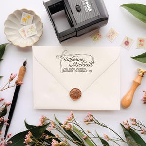 Könnte beinhalten: Ein cremefarbener Umschlag mit einem schwarzen Absenderstempel mit der Aufschrift Katherine Michaels, 7023 Egret Landing, Monroe, Louisiana 71203, versiegelt mit einem kupferfarbenen Wachssiegel und einem schwarzen Selbstfärbestempel.