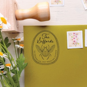 Könnte beinhalten: Ein Holzstempel mit Griff, platziert über einem olivgrünen Umschlag. Der Stempelabdruck zeigt einen ovalen Rand, eine Käfer-Illustration, den Text The Saffords sowie die Adresse 123 Presidio Ave, Half Moon Bay, CA 94019.