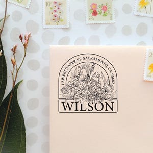 Peut inclure: Une empreinte de tampon à l'encre noire sur une enveloppe crème claire représentant un bouquet floral dans un cadre arqué. Le texte indique 1 SWEETWATER ST. SACRAMENTO, CA 95682 et WILSON. Le motif présente des dessins au trait de fleurs.