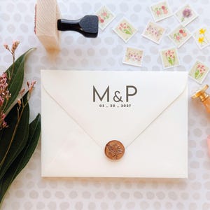 May include: Ivory envelope with the initials "M&P" and the date "03_20_2027" printed in gray. A bronze wax seal and floral stamps are scattered around the envelope. A wooden stamp and pink ribbon are also visible.
