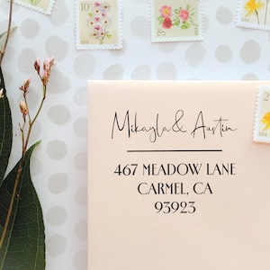 Peut inclure: Une enveloppe pêche clair avec un texte imprimé noir indiquant Mikayla & Austin, 467 Meadow Lane, Carmel, CA 93923. L'enveloppe est ornée de timbres postaux floraux vintage et placée à côté d'une branche de fleurs roses séchées.