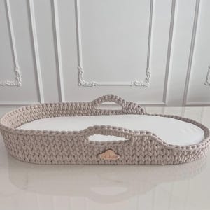 Può includere: Una culla per neonati all'uncinetto beige con un materasso bianco. La culla ha due maniglie e un'etichetta di legno con il nome "Elisa". Il design generale è di forma ovale.