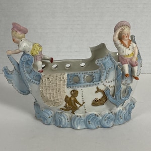 Maceta antigua de porcelana bizcochada con forma de barco, figura de marinero infantil y querubín náutico.