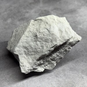 May include: A light gray, irregularly shaped rock with a rough, textured surface. The rock has a slightly angular form and appears to be a natural stone specimen. The background is a solid gray, providing contrast to the rock's color and texture.