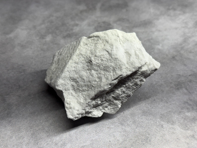 May include: A light gray, irregularly shaped rock with a rough, textured surface. The rock has a slightly angular form and appears to be a natural stone specimen. The background is a solid gray, providing contrast to the rock's color and texture.
