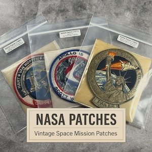May include: A collection of vintage NASA mission patches, including Apollo 15 and patches from 1971, 1984, and 1985. The patches are in clear plastic sleeves and feature colorful embroidered designs and text. The image includes a sign that says "NASA PATCHES" and "Vintage Space Mission Patches."
