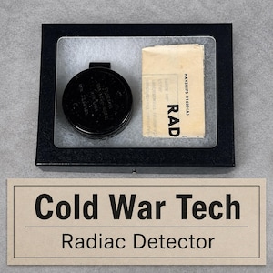 May include: A black circular radiation detector with embossed text in a black display box alongside a folded paper manual labeled NAYSHIPS 91609(A) RAD. Below the box is a rectangular card with the printed text Cold War Tech Radiac Detector.