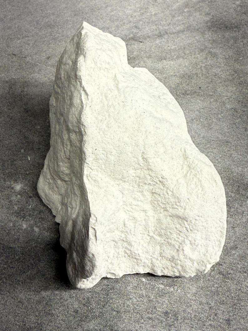May include: A large, irregularly shaped, off-white rock with a rough, textured surface. The rock has a natural, organic form, with visible ridges and variations in its surface. The background is a neutral gray.