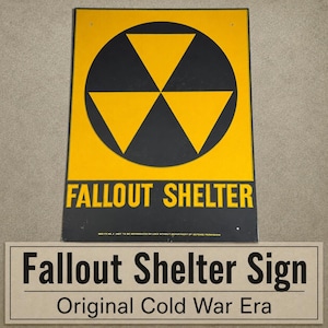May include: A rectangular yellow and black metal sign featuring a central circular radiation symbol with three triangular segments. The text FALLOUT SHELTER is printed in bold black capital letters below the symbol, with small text at the bottom.