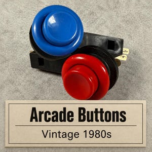 May include: Two circular arcade buttons, one blue and one red, each mounted on a black plastic housing with visible metal contact terminals. Below the buttons, a rectangular label features the black text Arcade Buttons and Vintage 1980s.