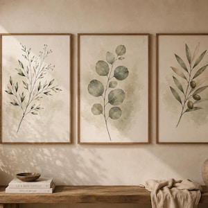 Op de afbeelding: Drie ingelijste botanische prints met aquarel illustraties van bladeren en takken in groen- en bruintinten. De prints worden weergegeven op een neutrale muur boven een houten bank met decoratieve items.
