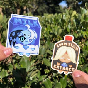 May include: Two stickers featuring cartoon characters. One is a blue milk carton with a character inside. The other is a vanilla extract bottle with a character. Both stickers have a white border and are held against a background of green foliage.