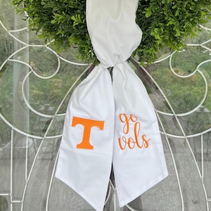 May include: A green, circular wreath with a white ribbon and orange lettering. The ribbon displays the letter "T" and the words "go vols". The wreath is hanging on a glass door.