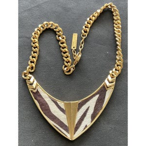 May include: Gold-tone necklace with a zebra print pendant. The pendant has a gold frame and a central vertical gold bar. The chain is a thick, gold curb chain. The necklace is on a dark gray surface.