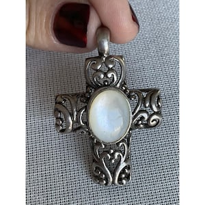May include: Silver-toned cross pendant with an oval, iridescent, white stone center. The cross features intricate, openwork filigree designs. The pendant is held by a hand with red nail polish.