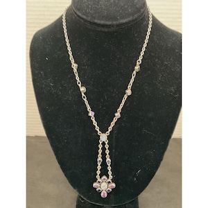 May include: Silver-toned necklace with a floral pendant. The necklace features a delicate chain with small, square and round accents. The pendant has a flower design with purple and white stones. The necklace is displayed on a black mannequin.