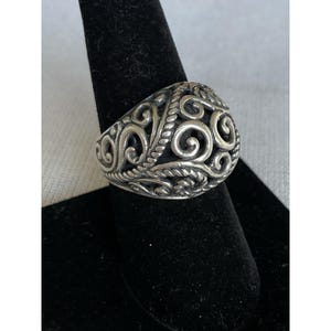 Puede incluir: Un anillo plateado con un diseño calado ornamentado con patrones arremolinados y detalles florales. El anillo se exhibe sobre un soporte negro, mostrando sus intrincados detalles y artesanía. El diseño del anillo recuerda a la joyería vintage o antigua.