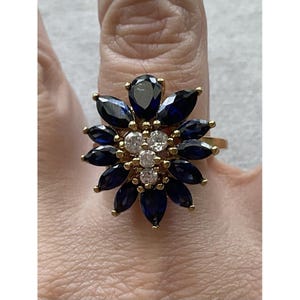 May include: A gold-toned ring featuring a cluster of dark blue marquise-cut stones arranged in a floral pattern, with a central grouping of four small, clear, round-cut stones. The setting is metallic gold, and the design creates a starburst effect.