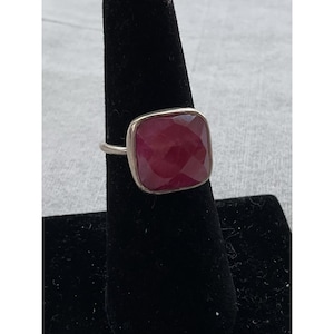 May include: A silver ring with a large, square, faceted, deep red gemstone. The gemstone is set in a silver bezel and has a polished finish. The ring is displayed on a black velvet ring holder.