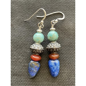 May include: A pair of handcrafted earrings with silver hooks. Each earring features a turquoise bead, a silver filigree bead, a red-orange bead, and a blue teardrop-shaped bead. The earrings are displayed against a textured gray background.