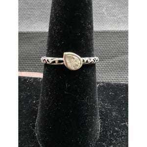 May include: A silver ring with a pear-shaped, light-colored gemstone. The band features an ornate, swirling design. The ring is displayed on a black velvet surface against a gray and white striped background.