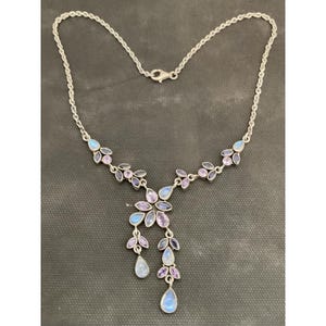 May include: A silver necklace with a floral design featuring blue, purple, and clear gemstones. The necklace has a delicate chain and a pendant with teardrop-shaped stones. The gemstones are set in a floral pattern.