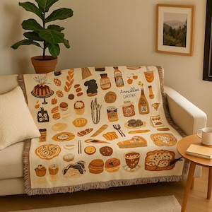 Puede incluir: Una manta beige con un patrón repetido de ilustraciones de alimentos, incluyendo pasteles, pan y botellas. La manta tiene un borde con flecos y las palabras "Annailles DRINK" están impresas en la tela. La manta está sobre un sofá blanco.