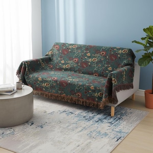 Könnte beinhalten: Eine grüne Decke mit floralem Muster, die über einem weißen Sofa drapiert ist. Die Decke hat ein sich wiederholendes Design aus roten und gelben Blumen. Der Überwurf hat einen braunen Fransenbesatz an den Kanten. Ein Couchtisch und ein Teppich befinden sich im Vordergrund.