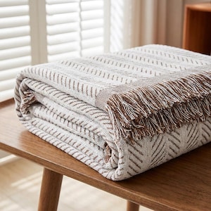 May include: A folded throw blanket featuring a cream and brown herringbone pattern. The textile has a woven texture with a decorative fringe edge in brown and cream tones along the side, showcasing a consistent geometric design throughout.