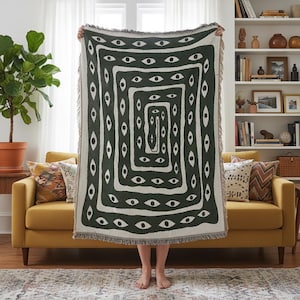 May include: A dark green and cream-colored woven throw blanket with a repeating eye pattern. The blanket has a rectangular design with a fringe border. The throw is held up in front of a yellow sofa and a bookshelf.