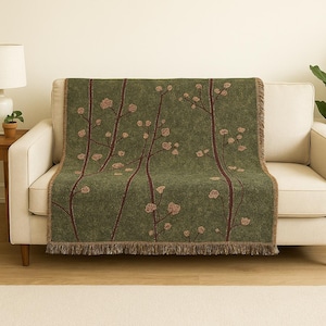 May include: A green throw blanket with a floral pattern draped over a beige sofa. The blanket features a repeating design of brown branches and peach-coloured blossoms. The edges are finished with a brown fringe.