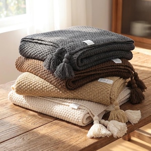 Könnte beinhalten: Ein Stapel aus vier gefalteten Waffelstrickdecken in Anthrazit, Dunkelbraun, Hellbraun und Creme. Jede Decke hat ein strukturiertes Gittermuster und ist mit passenden dekorativen Quasten versehen, die mit kleinen Holzperlen an den Ecken befestigt sind.