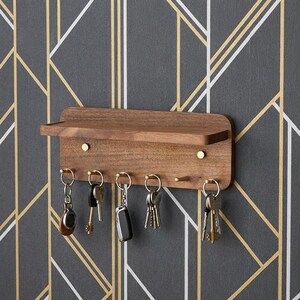 May include: A rectangular wall-mounted wooden key holder with a small top shelf and five brass-colored hooks. Several sets of metal keys with various keychains are hanging from the hooks. The unit features a smooth, natural wood grain finish.