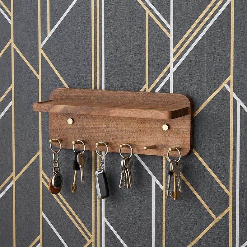May include: A rectangular wall-mounted wooden key holder with a small top shelf and five brass-colored hooks. Several sets of metal keys with various keychains are hanging from the hooks. The unit features a smooth, natural wood grain finish.
