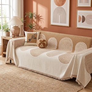 May include: A beige and tan sofa throw blanket featuring a repeating pattern of arched lines. The rectangular textile has a fringed edge and covers the seat and back of a sofa. A square throw pillow with a line-art face design sits on the sofa.