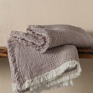 Peut inclure: Un plaid mauve plié présentant un motif gaufré texturé et de longues franges blanches sur les bords. Le tissu a un aspect doux et tactile avec une structure en grille cohérente sur toute la surface de ce textile décoratif pour la maison.