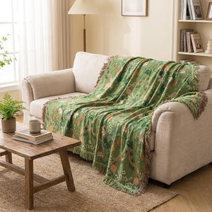 May include: A green woven throw blanket featuring a detailed botanical pattern with orange, white, and yellow flowers and green foliage. The blanket has a textured surface and is finished with decorative fringe along the edges, draped over a sofa.