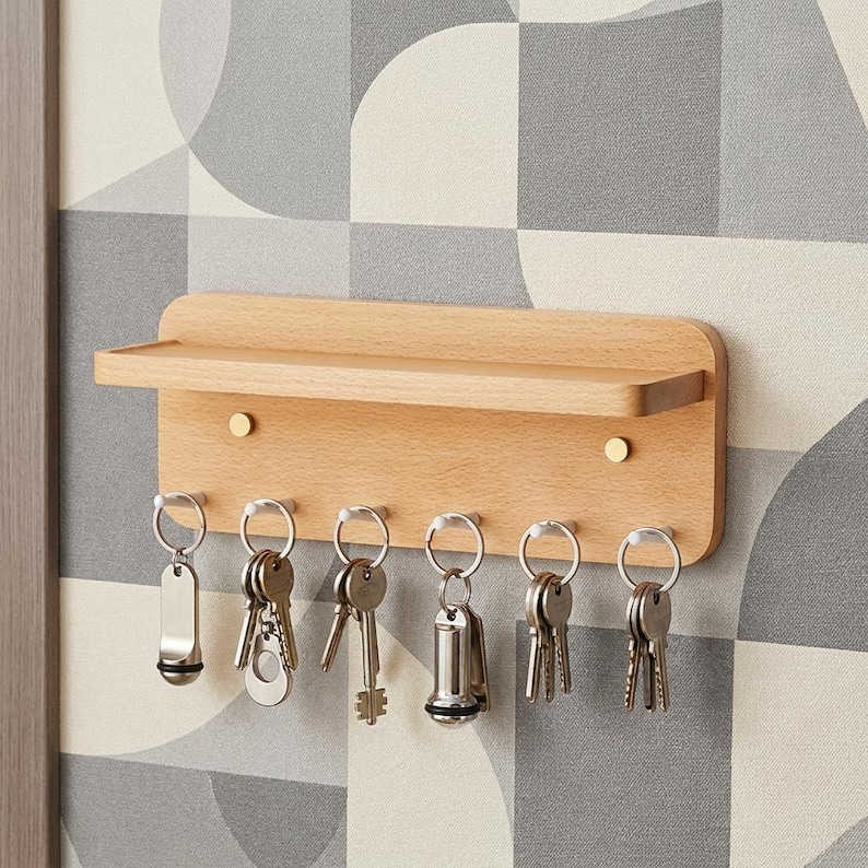 May include: A light-toned wooden wall-mounted key holder featuring a horizontal shelf at the top and six silver-colored metal hooks. Each hook holds a silver metal keyring with various keys attached. Two small gold-colored circular accents are visible.
