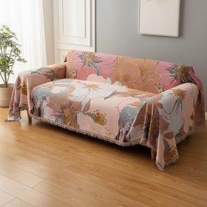 Puede incluir: Una funda de sofá con estampado floral en tonos rosa, melocotón y azul. La funda está colocada sobre un sofá con patas de madera y tiene bordes con flecos. El diseño presenta flores grandes y estilizadas.