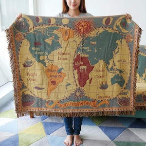 Può includere: Una coperta beige con un design a mappa del mondo vintage. La mappa presenta continenti in tonalità di verde, arancione e rosso, con nomi di oceani in marrone. La coperta ha un bordo con frange e le parole "AROUND WORLD" sono stampate in basso.