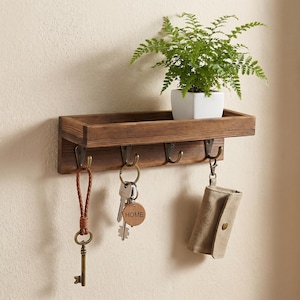 May include: A dark brown wooden wall-mounted shelf with four metal hooks. A green fern in a white pot sits on the shelf. Hanging from the hooks are a braided leather key fob with a metal key, a key ring with a wooden tag labelled HOME, and a beige pouch.