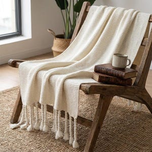 May include: A cream-coloured knit throw blanket with a textured pattern and long, twisted fringe tassels at the bottom edge. The blanket is draped over a wooden chair, with two stacked vintage books and a ceramic mug resting on the fabric surface.