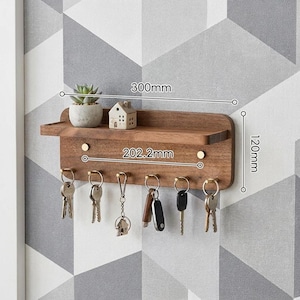 May include: A wall-mounted wooden key holder shelf with six brass hooks holding various keys. The shelf features a small succulent in a pot and a miniature house figurine. Dimensions are marked as 300mm wide, 120mm high, and 202.2mm between hooks.