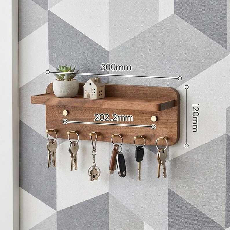 May include: A wall-mounted wooden key holder shelf with six brass hooks holding various keys. The shelf features a small succulent in a pot and a miniature house figurine. Dimensions are marked as 300mm wide, 120mm high, and 202.2mm between hooks.