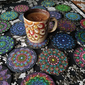 May include: A close-up of a cup of coffee on a coaster with a mandala design. The coaster is surrounded by other coasters with different mandala designs. The coasters are all black with colorful mandala designs.