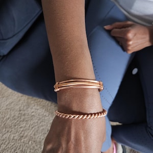 May include: Two copper bracelets are displayed on an arm. The top bracelet is a thick band with a wrapped wire design. The bottom bracelet has a twisted rope design. The bracelets are a warm, reddish-brown color.