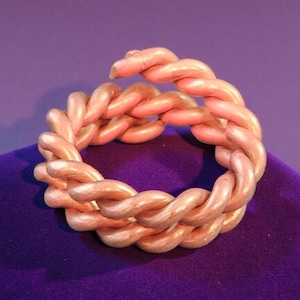 May include: A braided ring in a soft peach color, resting on a purple surface. The ring's design features intertwined strands, creating a textured and dimensional appearance. The ring is a unique piece of jewelry.