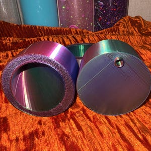 Puede incluir: Dos objetos cilíndricos iridiscentes con un borde texturizado y una parte superior plana y acanalada. Los colores cambian de morado a verde. Los objetos están sobre una superficie naranja texturizada.