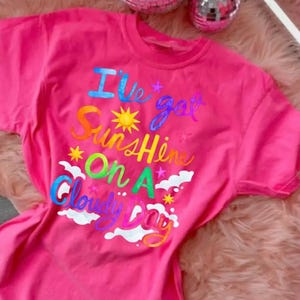 Puede incluir: Una camiseta rosa brillante con la frase "I've got Sunshine On A Cloudy Day" en letras coloridas y caprichosas. El diseño incluye un sol, estrellas y nubes. La camiseta se muestra sobre una superficie rosa y esponjosa.